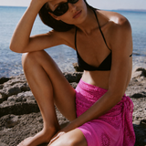 Pembe blok baskı desenli plaj örtüsü, siyah bikini ve güneş gözlüğüyle kaya üzerinde oturan kadın