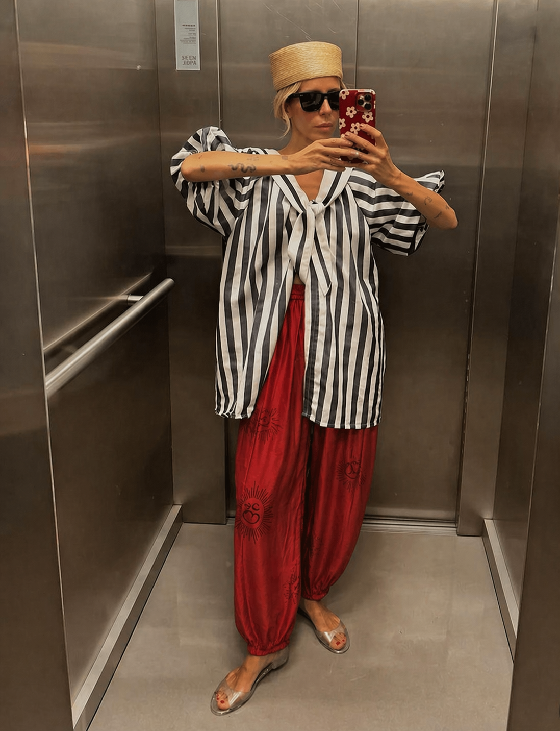 Kadın, siyah beyaz çizgili bluz ve kırmızı blok baskı pantolon giyerek asansörde ayna selfiesi çekiyor.